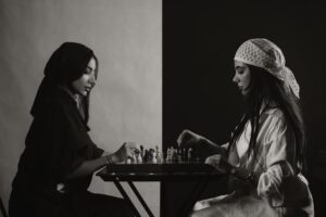 Two women play chess at a table in an artistic black and white setting.