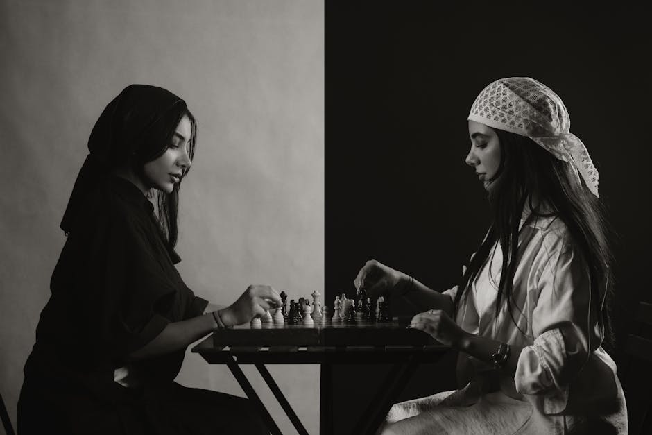 Two women play chess at a table in an artistic black and white setting.