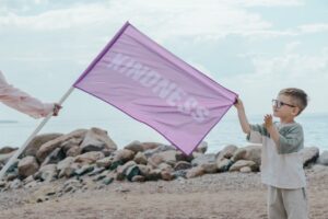 A young boy holding a pink flag with 'Kindness' on a rocky beach setting.