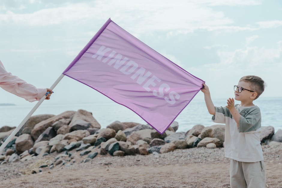 A young boy holding a pink flag with 'Kindness' on a rocky beach setting.