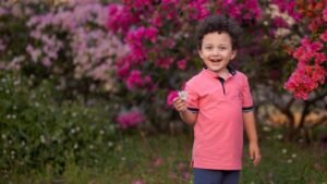 Young boy in pink polo shirt holding a flower, surrounded by vibrant spring blooms.