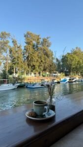 Enjoy a serene river view with coffee, boats, and lush trees in the warm daylight.