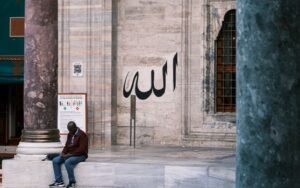 A man sits near a mosque entrance with Arabic calligraphy on the stone wall.
